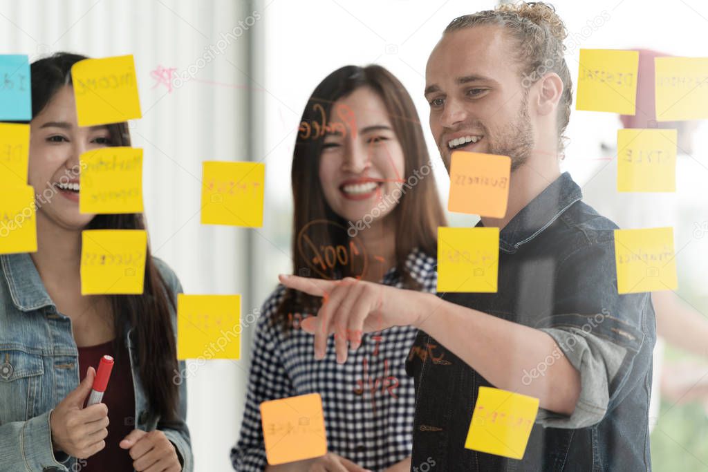 Group of young successful creative multiethnic team smile and brainstorm on project together in modern office with post note or sticky note. Caucasian man point on glass wall sharing idea.