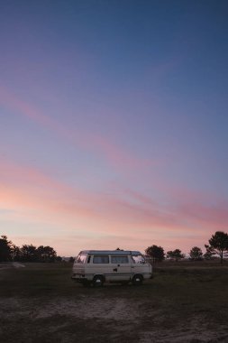Gün batımında park yeri manzarasında logoları olmayan beyaz, orta büyüklükte bir hippi minibüsü. Macera, açık hava eğlencesi, doğa maceraları, tatil. 