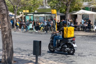 Moto Scooter Outdoor 'a servis yapan teslimatçı elinde yemek servisi olan sarı termal çantasıyla Coronavirus ya da Cvid-19 yüzünden daha fazla korunmak için restoran yemekleri getiriyor. Yemek Teslimatı