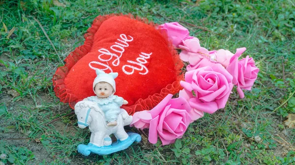 Romantic decoration with a red heart-shaped cushion reading I Love You, surrounded by pink roses and a baby doll figurine on grass. Perfect for Valentines Day, love, romance, wedding, and special celebration themes.