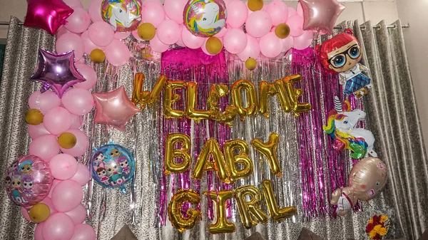 Festive decoration setup for a baby girl celebration with pink balloons, golden foil letters saying Welcome Baby Girl, unicorn and cartoon balloons, shiny backdrop, and colourful party atmosphere. 