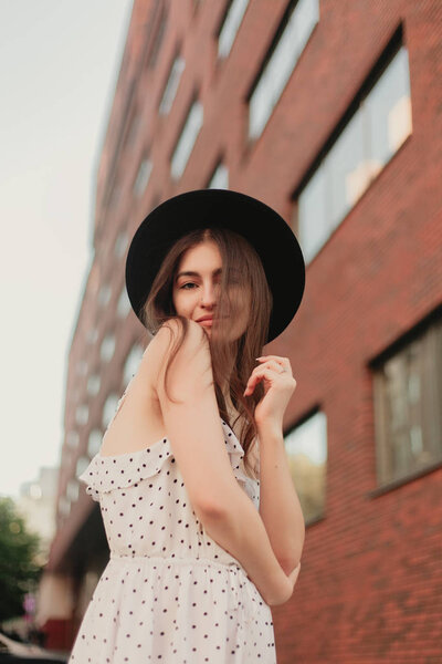 young teenage girl in a white dress and hat. style of the big city. Sunset sky and rays of the sun. Natural beauty pretty woman in black fedora hipster hat, on warm summer evening sunset in big city