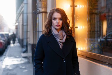 Hevesli gülümseyen kız ile parlak güneş ışığı altında şehirde poz bukleler. Yakın çekim açık harika gülümseme ve büyük mavi gözleri ile moda sonbahar siperde büyüleyici Bayan portresi.