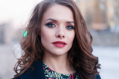 Hevesli gülümseyen kız ile parlak güneş ışığı altında şehirde poz bukleler. Yakın çekim açık harika gülümseme ve büyük mavi gözleri ile moda sonbahar siperde büyüleyici Bayan portresi.