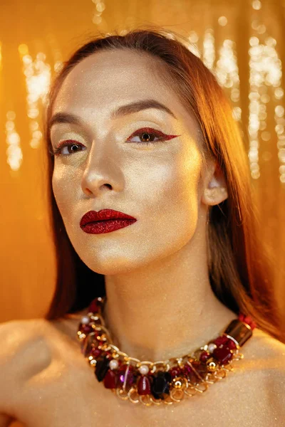 Magic Girl Portrait in Gold. Golden Makeup, close-up portrait in studio shot, color. Beauty Model Girl with perfect bright make-up, red lips, golden maroon jewellery. Sexy lady makeup Holiday party.