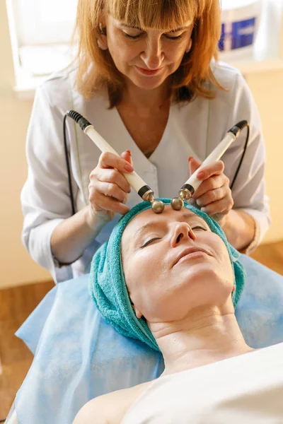 people, beauty, spa, cosmetology and technology concept - Cosmetologist makes the procedure microcurrent therapy of the facial skin of a beautiful, young woman in a beauty salon