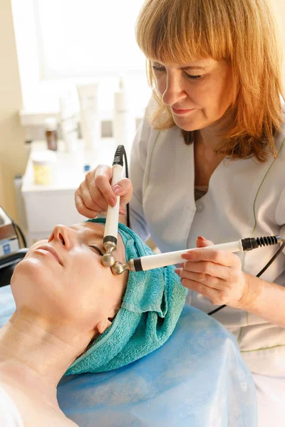 people, beauty, spa, cosmetology and technology concept - Cosmetologist makes the procedure microcurrent therapy of the facial skin of a beautiful, young woman in a beauty salon