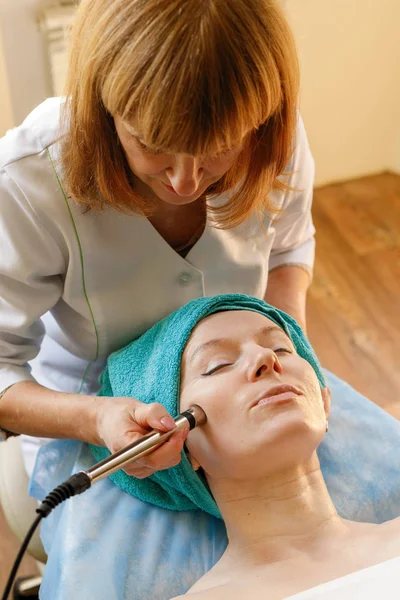 people, beauty, spa, cosmetology and technology concept - Cosmetologist makes the procedure microcurrent therapy of the facial skin of a beautiful, young woman in a beauty salon