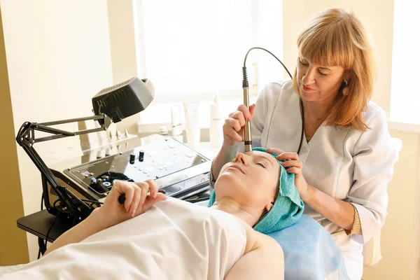 people, beauty, spa, cosmetology and technology concept - Cosmetologist makes the procedure microcurrent therapy of the facial skin of a beautiful, young woman in a beauty salon