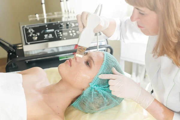 people, beauty, spa, cosmetology and technology concept - The cosmetologist makes the procedure ultrasonic face peeling of the facial skin of a beautiful, young woman in a beauty salon