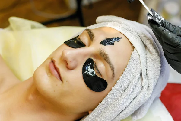 people, beauty, spa, cosmetology and technology concept - Spa salon. The cosmetologist puts a mask on the face of a young man.