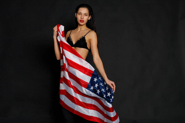 Portrait of proud female athlete wrapped in American Flag against black background. Muscular young woman looking confidently at camera.