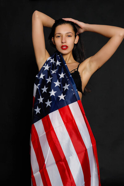 Portrait of proud female athlete wrapped in American Flag against black background. Muscular young woman looking confidently at camera.