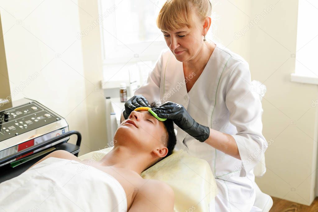 people, beauty, spa, cosmetology and technology concept - The cosmetologist makes the procedure ultrasonic face peeling of the facial skin of a beautiful, young man in a beauty salon
