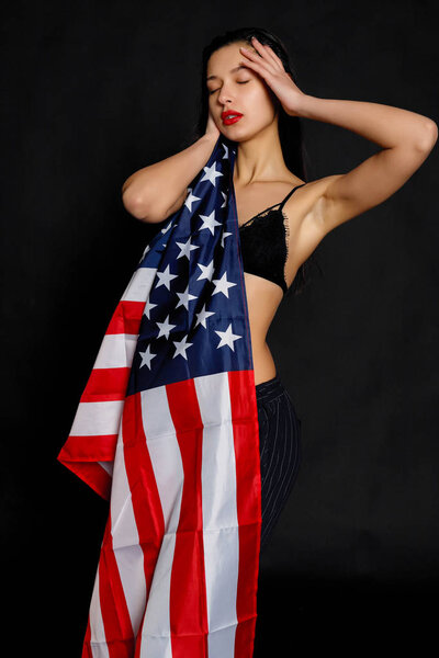 Portrait of proud female athlete wrapped in American Flag against black background. Muscular young woman looking confidently at camera.