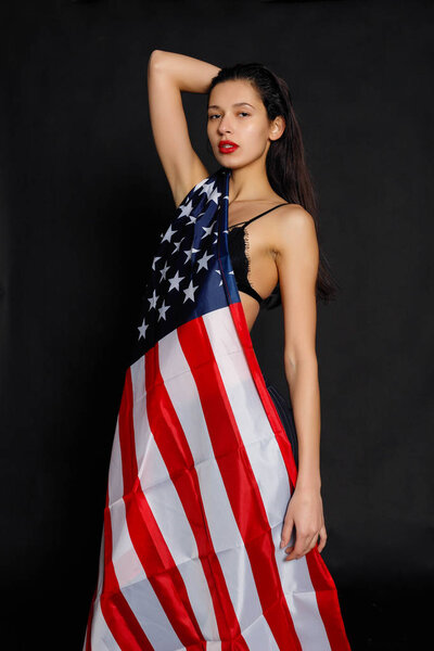 Portrait of proud female athlete wrapped in American Flag against black background. Muscular young woman looking confidently at camera.