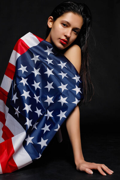 Portrait of proud female athlete wrapped in American Flag against black background. Muscular young woman looking confidently at camera.