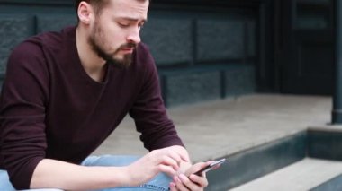 Portre adam oturan sokak şehir merkezinde yana doğru gülümseme görünümlü telefon kullanın. Mutlu Gülümseyen Çekici Adam Sokakta Yürürken. İlgiyle Akıllı Telefonunda Yazma.