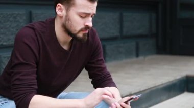 Portre adam oturan sokak şehir merkezinde yana doğru gülümseme görünümlü telefon kullanın. Mutlu Gülümseyen Çekici Adam Sokakta Yürürken. İlgiyle Akıllı Telefonunda Yazma.