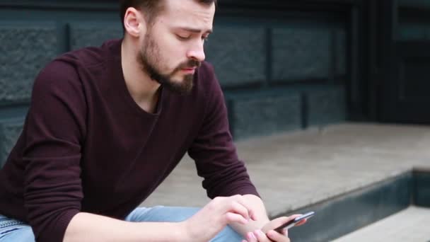 肖像人坐在使用手机侧视侧身微笑在街道市中心 快乐微笑有吸引力的男人走在街上 有兴趣在智能手机上打字 — 图库视频影像