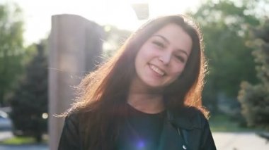 Şehrin gün batımının arka planında güzel bir genç kadının portresi. Şehirde sokakta yürüyen deri ceketli çekici kız, güneş bahar güneşini parlatıyor. Yavaş çekim