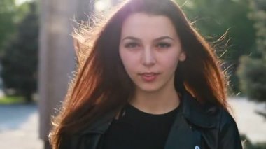 Şehrin gün batımının arka planında güzel bir genç kadının portresi. Şehirde sokakta yürüyen deri ceketli çekici kız, güneş bahar güneşini parlatıyor. Yavaş çekim