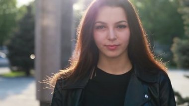 Şehrin gün batımının arka planında güzel bir genç kadının portresi. Şehirde sokakta yürüyen deri ceketli çekici kız, güneş bahar güneşini parlatıyor. Yavaş çekim