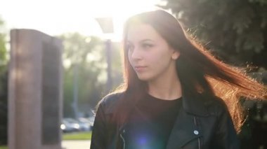 Şehrin gün batımının arka planında güzel bir genç kadının portresi. Şehirde sokakta yürüyen deri ceketli çekici kız, güneş bahar güneşini parlatıyor. Yavaş çekim