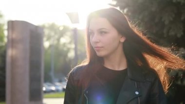 Şehrin gün batımının arka planında güzel bir genç kadının portresi. Şehirde sokakta yürüyen deri ceketli çekici kız, güneş bahar güneşini parlatıyor. Yavaş çekim