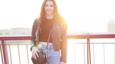 Şehrin gün batımının arka planında güzel bir genç kadının portresi. Şehirde sokakta yürüyen deri ceketli çekici kız, güneş bahar güneşini parlatıyor. Yavaş çekim