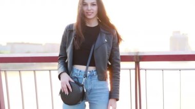 Şehrin gün batımının arka planında güzel bir genç kadının portresi. Şehirde sokakta yürüyen deri ceketli çekici kız, güneş bahar güneşini parlatıyor. Yavaş çekim