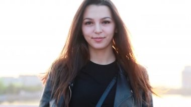 Şehrin gün batımının arka planında güzel bir genç kadının portresi. Şehirde sokakta yürüyen deri ceketli çekici kız, güneş bahar güneşini parlatıyor. Yavaş çekim