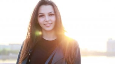 Şehrin gün batımının arka planında güzel bir genç kadının portresi. Şehirde sokakta yürüyen deri ceketli çekici kız, güneş bahar güneşini parlatıyor. Yavaş çekim