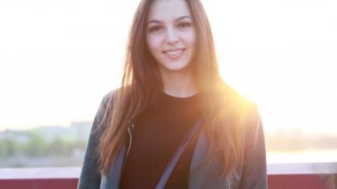 Şehrin gün batımının arka planında güzel bir genç kadının portresi. Şehirde sokakta yürüyen deri ceketli çekici kız, güneş bahar güneşini parlatıyor. Yavaş çekim