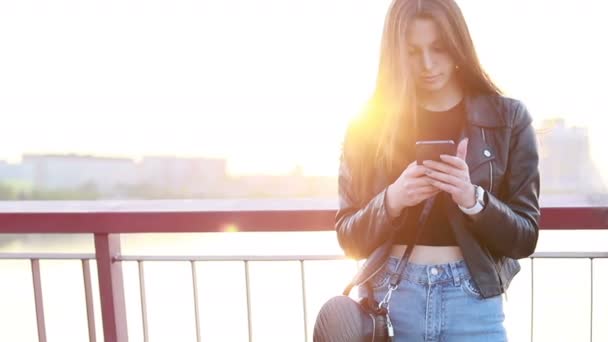 在城市日落的背景下 一个美丽的年轻女子的肖像 年轻时髦的女孩穿着皮夹克 并通过智能手机在社交网络上与朋友聊天 — 图库视频影像