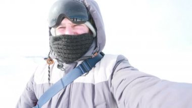 Selfi zamanı. Kapüşonlu, kar kayaklı ya da kayak ceketli mutlu gülümseyen bir adam bayırda durur ve güneşli bir kış günü eğlence için selfie çeker. hafif video, yüksek ağ anahtarı, az gösterilen