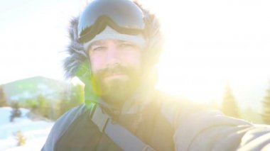 Selfi zamanı. Kapüşonlu, kar kayaklı ya da kayak ceketli mutlu gülümseyen bir adam bayırda durur ve güneşli bir kış günü eğlence için selfie çeker. hafif video, yüksek ağ anahtarı, az gösterilen