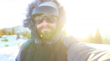 Selfi zamanı. Kapüşonlu, kar kayaklı ya da kayak ceketli mutlu gülümseyen bir adam bayırda durur ve güneşli bir kış günü eğlence için selfie çeker. hafif video, yüksek ağ anahtarı, az gösterilen