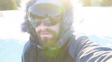 Selfi zamanı. Kapüşonlu, kar kayaklı ya da kayak ceketli mutlu gülümseyen bir adam bayırda durur ve güneşli bir kış günü eğlence için selfie çeker. hafif video, yüksek ağ anahtarı, az gösterilen