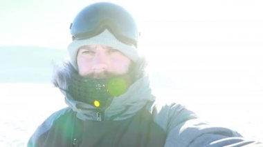 Selfi zamanı. Kapüşonlu, kar kayaklı ya da kayak ceketli mutlu gülümseyen bir adam bayırda durur ve güneşli bir kış günü eğlence için selfie çeker. hafif video, yüksek ağ anahtarı, az gösterilen