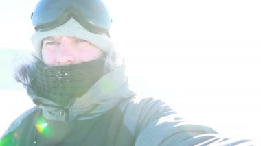 Selfi zamanı. Kapüşonlu, kar kayaklı ya da kayak ceketli mutlu gülümseyen bir adam bayırda durur ve güneşli bir kış günü eğlence için selfie çeker. hafif video, yüksek ağ anahtarı, az gösterilen