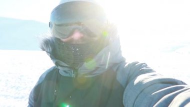 Selfi zamanı. Kapüşonlu, kar kayaklı ya da kayak ceketli mutlu gülümseyen bir adam bayırda durur ve güneşli bir kış günü eğlence için selfie çeker. hafif video, yüksek ağ anahtarı, az gösterilen