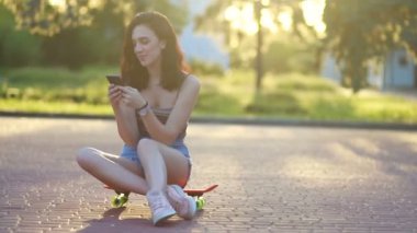 Selfie zamanı. Yerde oturan çekici kız akıllı telefonunda özçekim fotoğrafları çekiyor. Günbatımında kaykayın başında gülümseyen genç bir kızın portresi. Yavaş çekim
