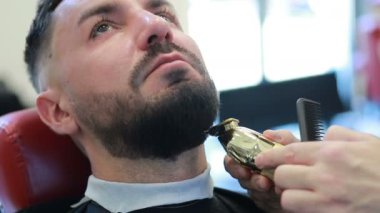 Saç şekillendirme işlemi. Genç sakallı bir adamın saçını kurutan berberin yakın çekimi. Berber dükkanındaki genç adam koltukta oturuyor. Berber saçını budayıcı ile kesiyor.