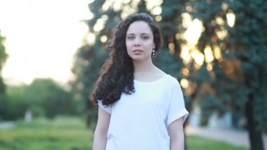 Günışığı, kıvırcık saçlı, gülümseyen genç kadın, kameraya bak, gülümse, parkta gülümse, gülümse, mutlu, yavaş çekimde yaz güneşi, gün batımına bak.