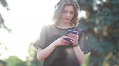 Güzel bir bayan model şehirde duruyor ve cep telefonuyla müzik dinliyor. Modern kulaklıklar kullanarak. Screen 'e yazıp popüler şarkıları seçmek. Çekici görünüş ve şık kıyafet..