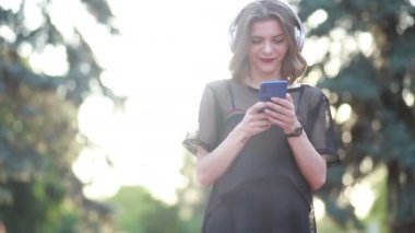 Güzel bir bayan model şehirde duruyor ve cep telefonuyla müzik dinliyor. Modern kulaklıklar kullanarak. Screen 'e yazıp popüler şarkıları seçmek. Çekici görünüş ve şık kıyafet..