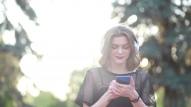 Güzel bir bayan model şehirde duruyor ve cep telefonuyla müzik dinliyor. Modern kulaklıklar kullanarak. Screen 'e yazıp popüler şarkıları seçmek. Çekici görünüş ve şık kıyafet..