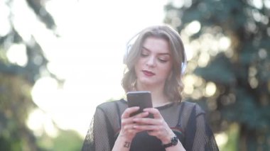 Güzel bir bayan model şehirde duruyor ve cep telefonuyla müzik dinliyor. Modern kulaklıklar kullanarak. Screen 'e yazıp popüler şarkıları seçmek. Çekici görünüş ve şık kıyafet..