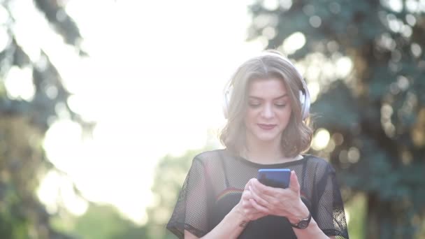 美丽的女模特站在镇上 用她的手机听音乐 使用现代耳机 在屏幕上打字 选择流行歌曲 迷人的外表和时髦的服装 — 图库视频影像